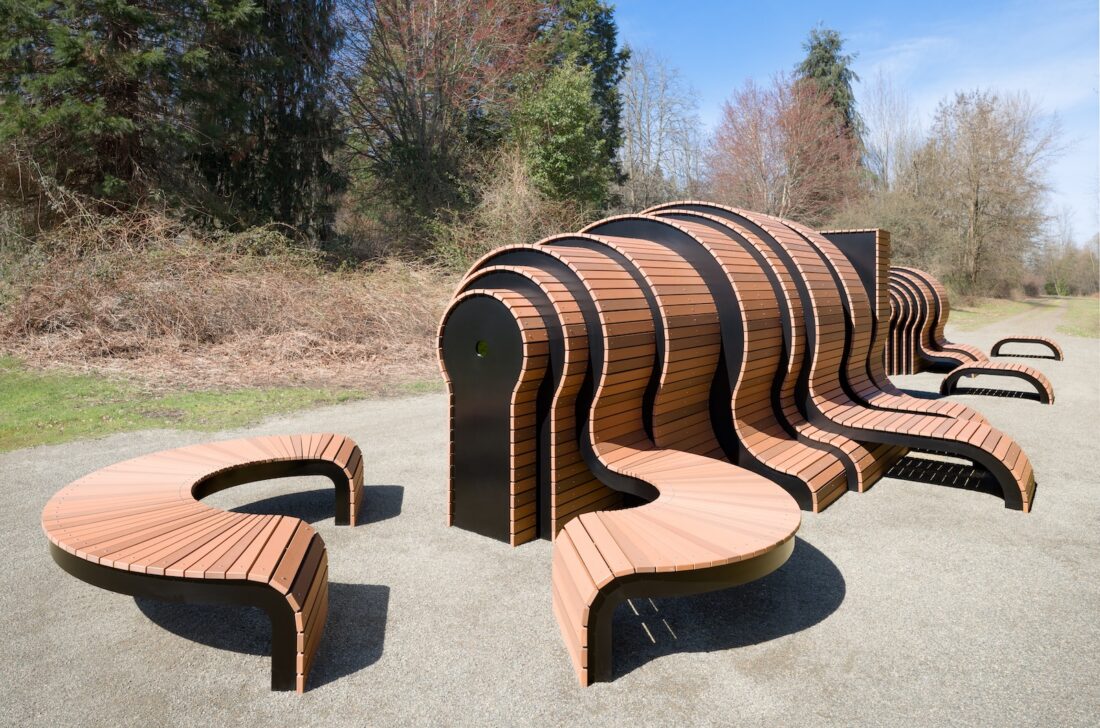 A uniquely designed outdoor bench features curved, wave-like wooden slats forming sculptural seating in a park setting, surrounded by trees and bare shrubs under a clear sky.
