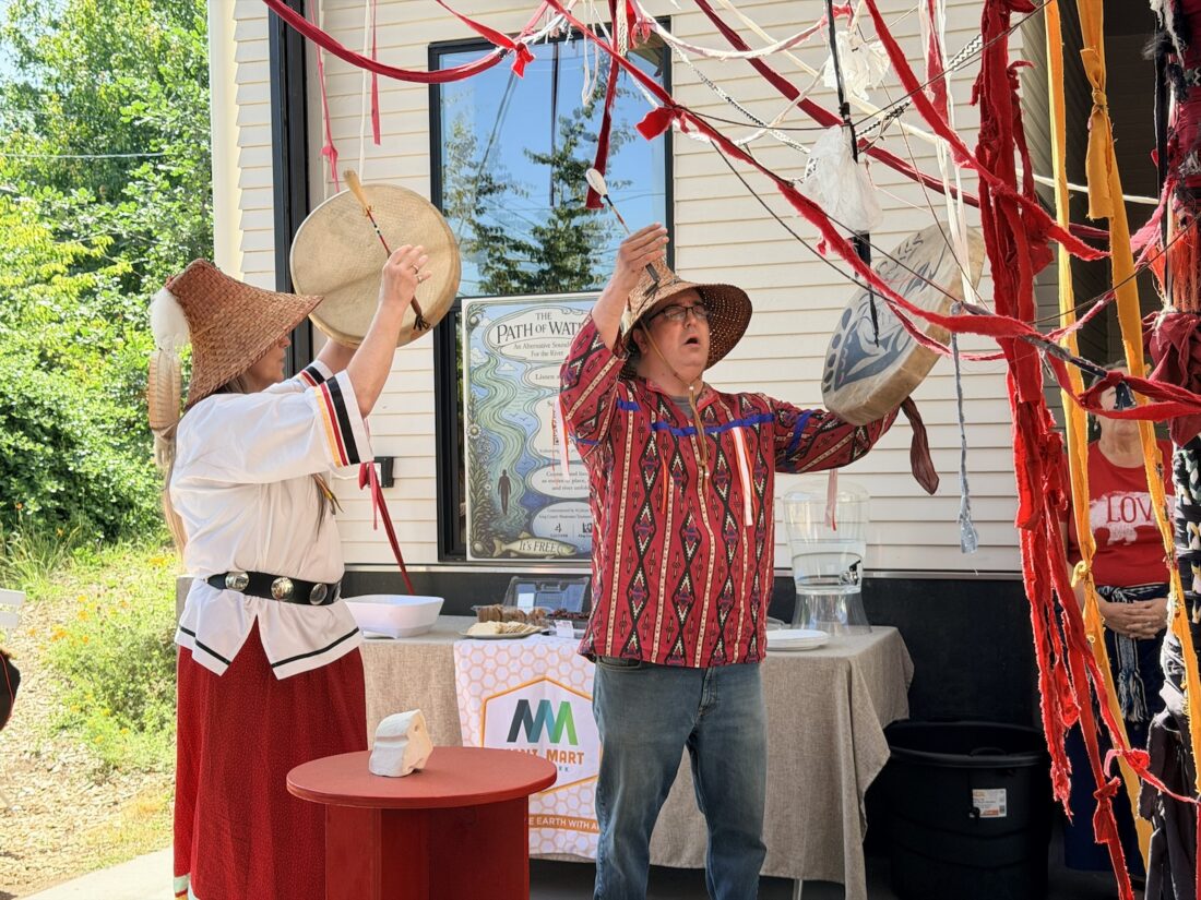 Two people in woven hats and traditional clothing drum and gesture near red ribbons and fabric outside a building, with greenery in the background and a table with items behind them.