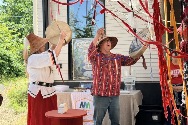 Two people in woven hats and traditional clothing drum and gesture near red ribbons and fabric outside a building, with greenery in the background and a table with items behind them.