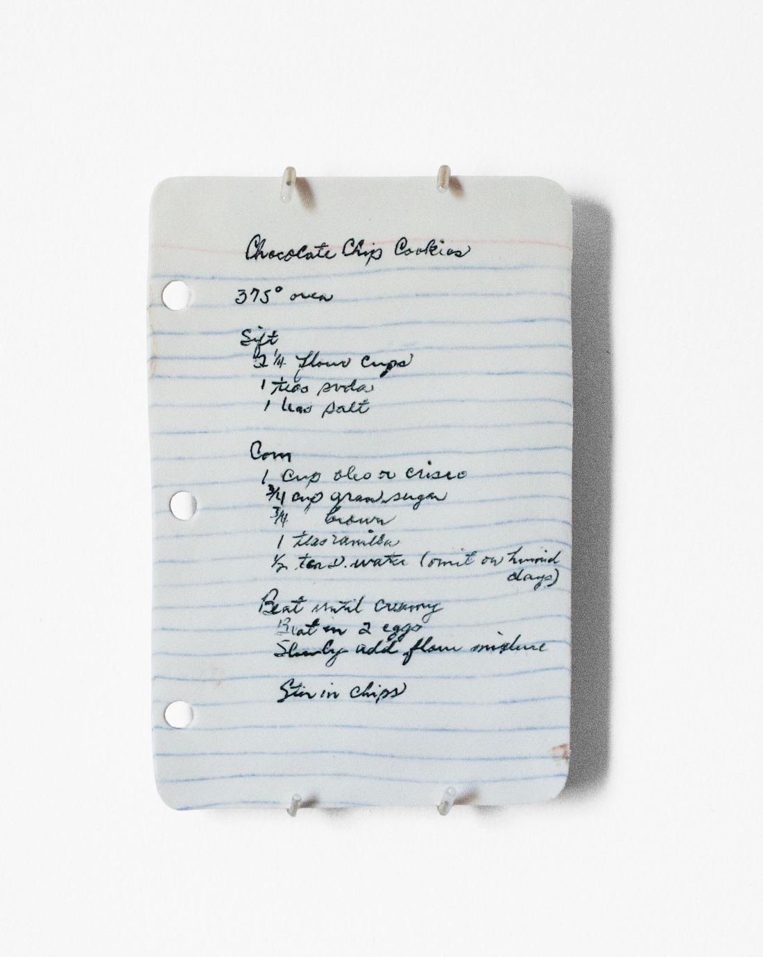 A handwritten chocolate chip cookie recipe on a piece of lined paper with three ring binder holes, written in blue ink and slightly tilted on a white background.