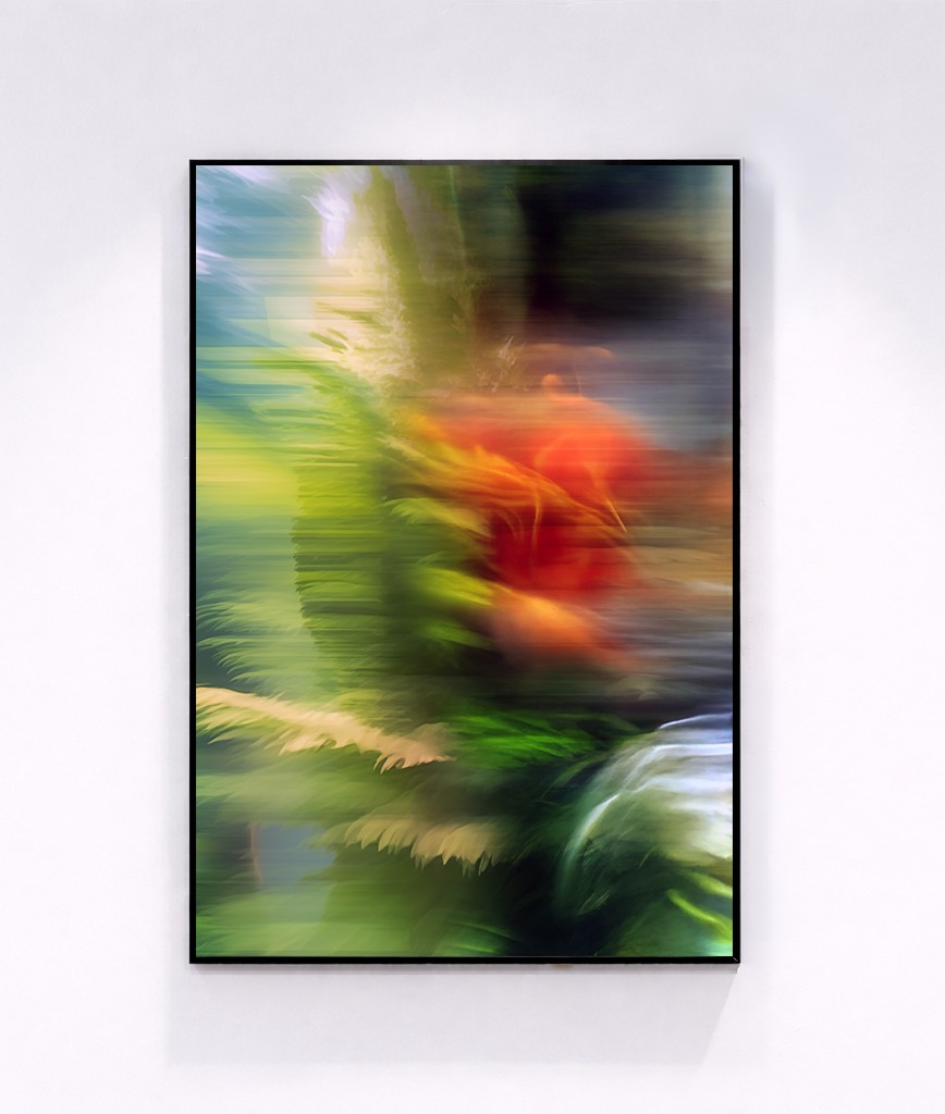 A framed abstract artwork featuring vibrant, blurred streaks of red, green, yellow, and blue, evoking the impression of flowers and foliage in motion, displayed against a plain white wall.