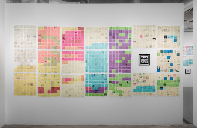 A gallery wall displays a grid of colorful papers—pink, orange, green, blue, and purple—arranged in large blocks. Two framed artworks are also hung among the papers. The floor and ceiling are visible.
