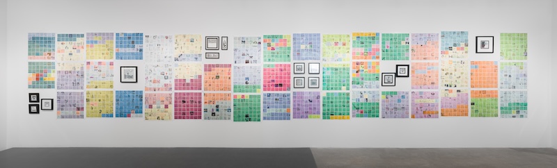 A gallery wall displays a large grid of colorful, square-filled panels arranged in rows, interspersed with several framed black-and-white drawings. The artwork covers a wide section of the white wall above a gray floor.
