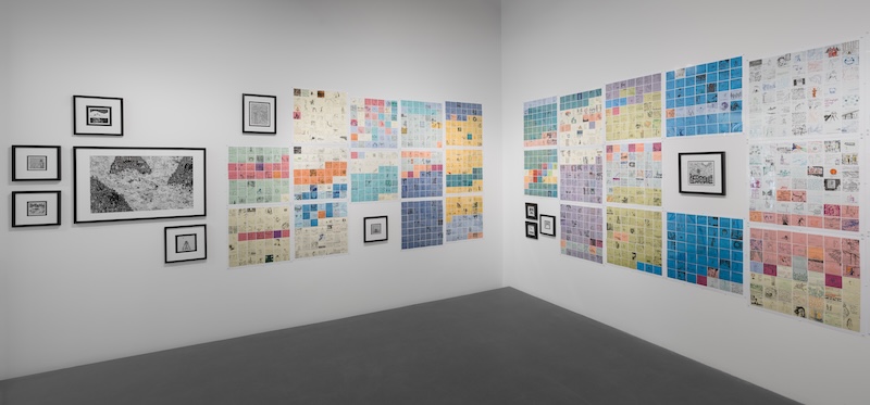 Art gallery corner with framed black and white prints on one wall and colorful grid-arranged works covering two adjacent walls, featuring varied images and text on pastel backgrounds. Gray floor and white walls.