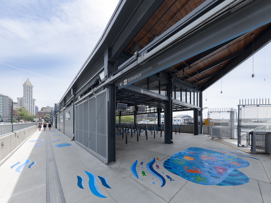 A waterfront ferry terminal entrance with blue aquatic-themed artwork on the pavement, metal railings, and a modern, open-roof structure. A city skyline and a tall building are visible in the background.