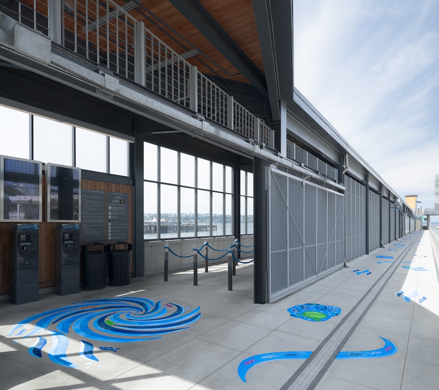 A modern outdoor walkway with large windows, interactive displays, and colorful blue and green spiral and wave patterns painted on the concrete floor under a partly cloudy sky.
