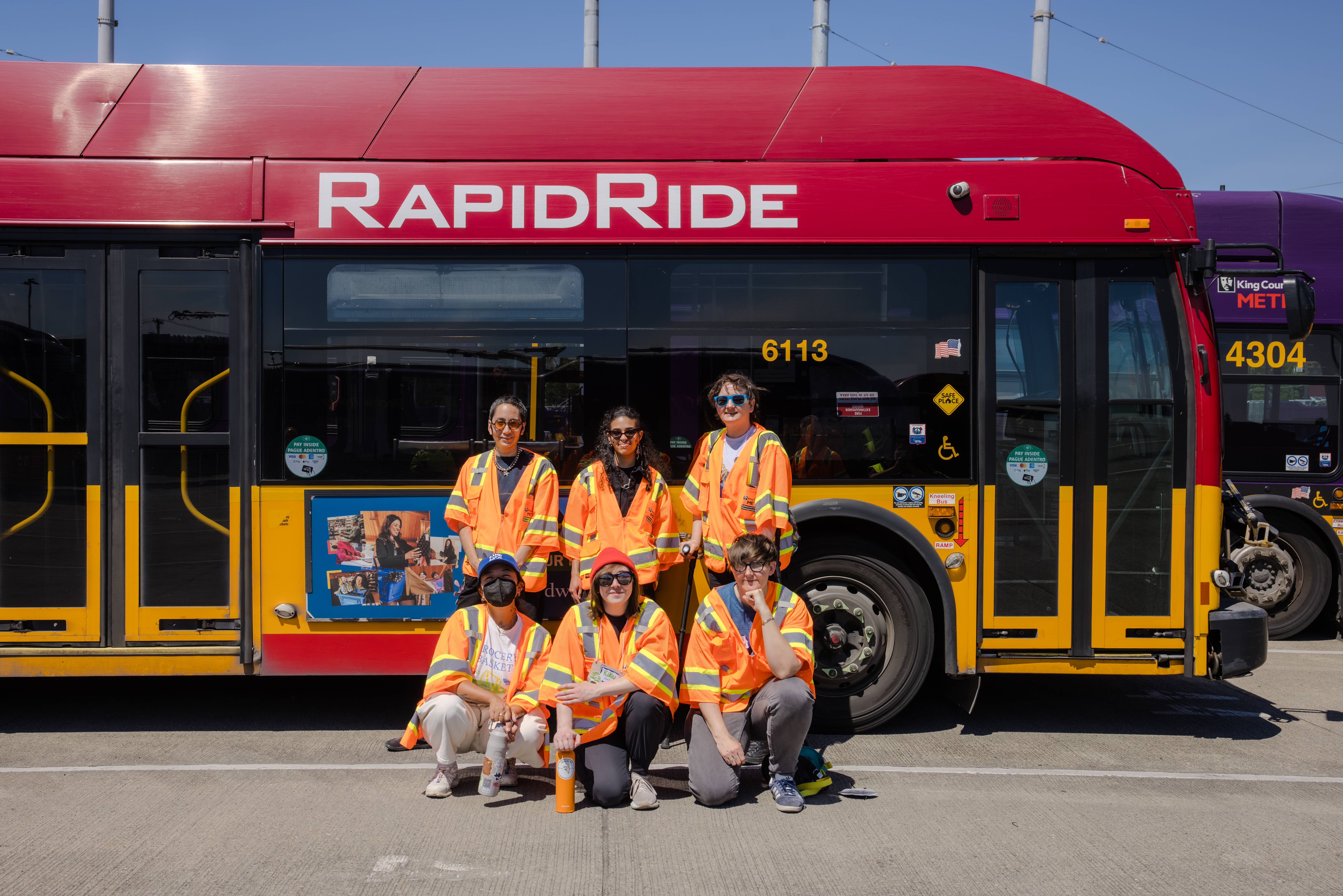 Six people wearing orange safety vests pose in front of a red and yellow RapidRide bus, with another bus partially visible in the background, on a sunny day.