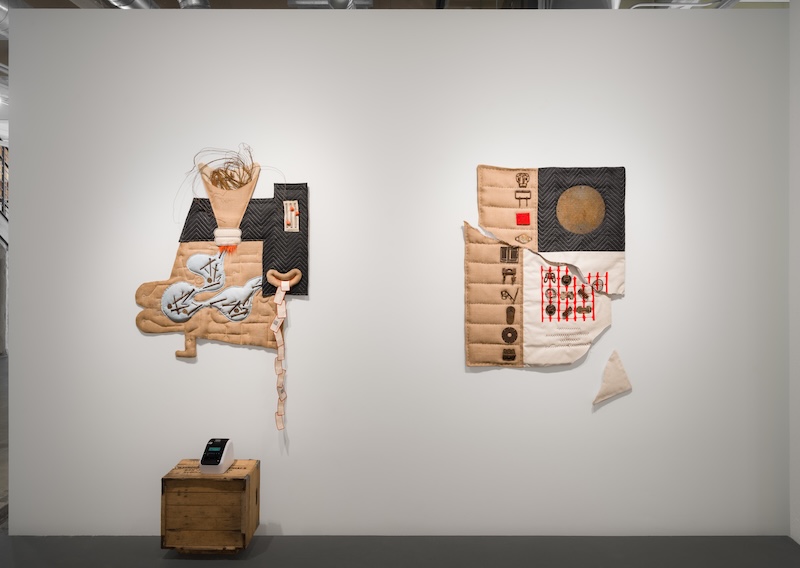 Two abstract textile artworks hang on a white gallery wall; each incorporates quilted, padded, and stitched fabric with various patterns. A small wooden crate with a device on top sits on the floor beneath the art.