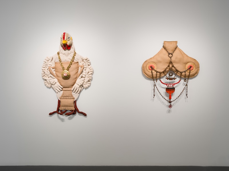 Two abstract mixed-media wall sculptures: one resembles a white chicken with red and yellow details, the other has a symmetrical, tan, mask-like form with beads, metal chains, and red accents, both mounted on a gray gallery wall.