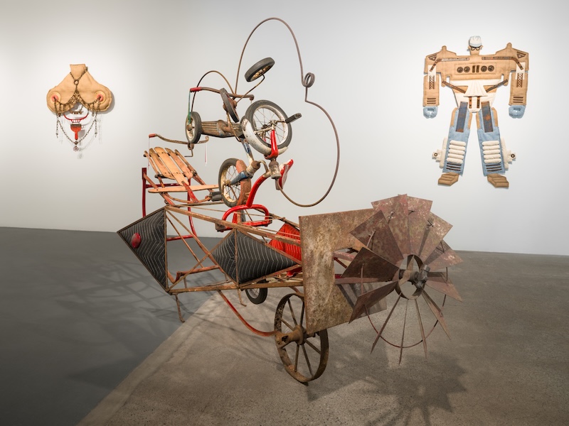A whimsical sculpture made from a tricycle and metal parts stands in a gallery, with two colorful, mask-like mixed-media artworks hanging on the white wall behind it.