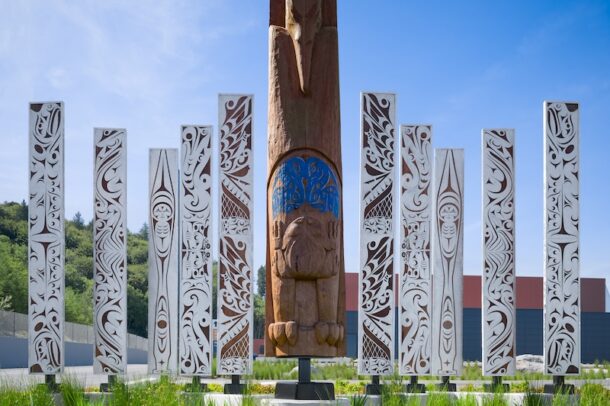 A tall wooden totem with carved figures stands surrounded by several white, intricately patterned pillars in an outdoor garden, with green grass and a clear blue sky.