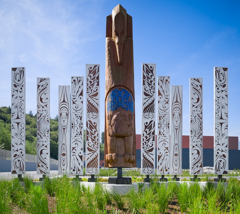 A tall wooden totem with carved figures stands surrounded by several white, intricately patterned pillars in an outdoor garden, with green grass and a clear blue sky.