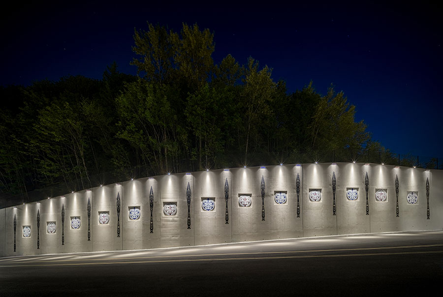 A well-lit concrete wall at night features evenly spaced illuminated art panels, with trees visible behind the wall and a road passing in front.