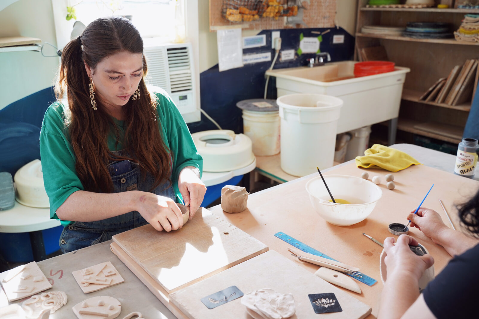 Two people work with clay at a pottery studio table, shaping pieces and using brushes and tools. Sunlight streams in, highlighting art supplies and ceramics around the workspace.