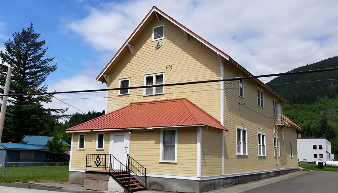 A yellow, two-story wooden building with a red metal roof and a Masonic square and compass symbol on the gable.