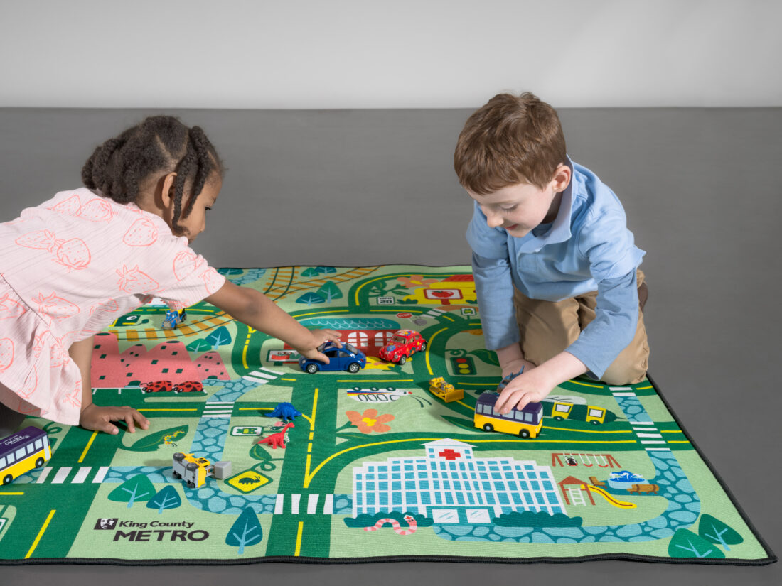 Two children play with toy vehicles on a King County Metro themed rug featuring a city map.