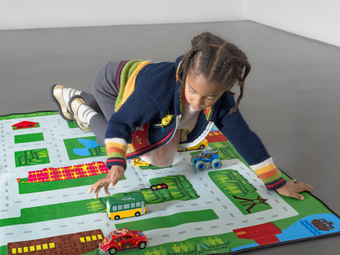 A child crawls on a play rug printed with a city street map, playing with a yellow and green toy bus and several other toy vehicles.
