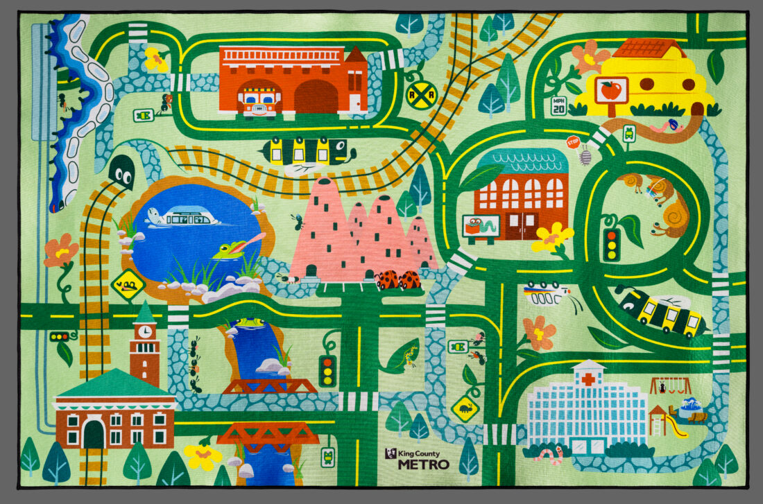 A colorful illustrated play rug featuring a map of a town with roads, train tracks, a lake, and various buildings including a station, hospital, and school, branded with the King County Metro logo.