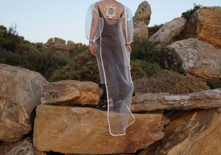 A person standing on a rocky hillside wearing a long, sheer white hooded garment with a prominent circular embroidered detail on the back.
