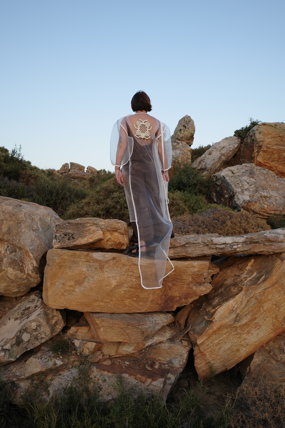 A person standing on a rocky hillside wearing a long, sheer white hooded garment with a prominent circular embroidered detail on the back.